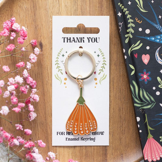 A floral enamel keyring, shaped like an orange flower petal, hangs on a Thank You card with leafy designs—a delightful teacher appreciation gift placed next to pink flowers and a black notebook with colourful floral patterns.