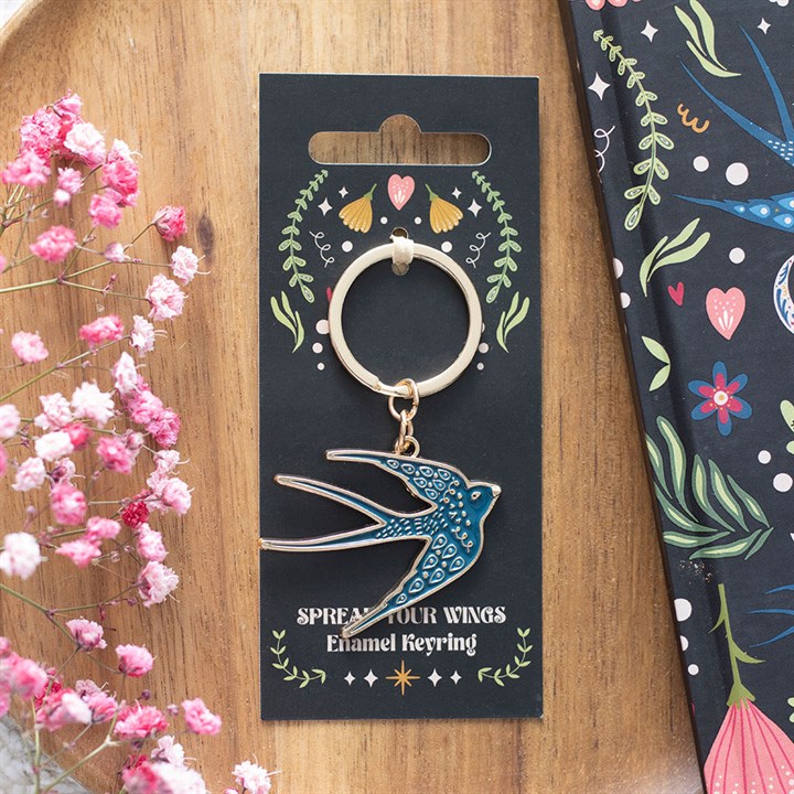 A blue swallow enamel keyring on black floral packaging featuring the words SPREAD YOUR WINGS, presented on wooden surface with pink babys breath—an ideal, charming gift for nature lovers.