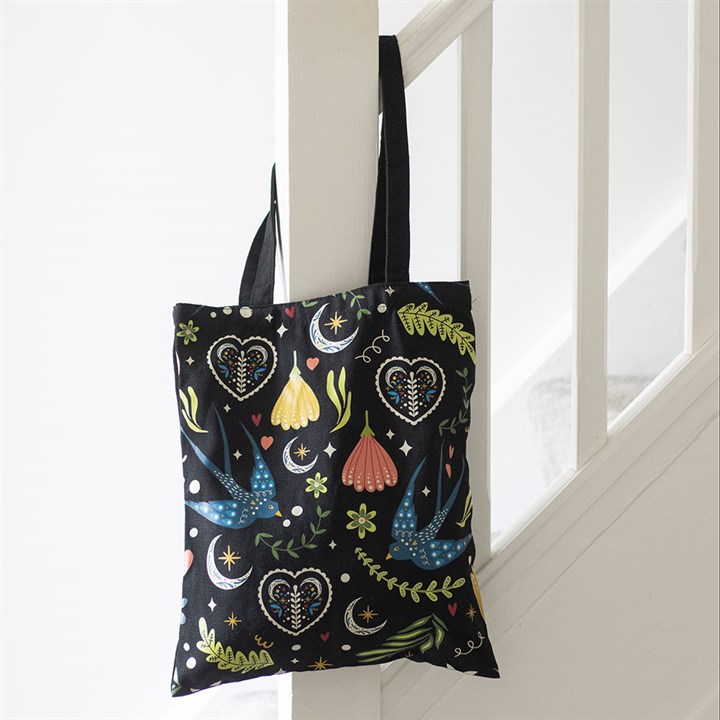 A black polycotton tote bag with colourful folk-art designs of birds, flowers, hearts, leaves, stars, and crescent moons hangs on a white stair bannister in a bright hallway.
