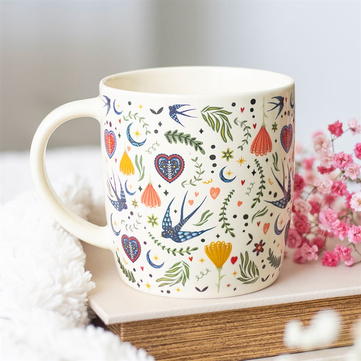 A cream-coloured ceramic mug with a handle, adorned with birds, hearts, flowers, leaves, and stars in blue, green, red, and yellow. The mug rests on a book next to pink flowers and a soft white cloth.