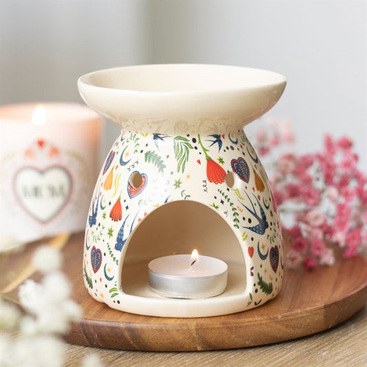 A decorative ceramic oil burner with colourful folk patterns holds a lit tealight inside. Wax melts or fragrance oil can be added, scenting the room as it rests near pink and white flowers and a blurred “MUM” candle.
