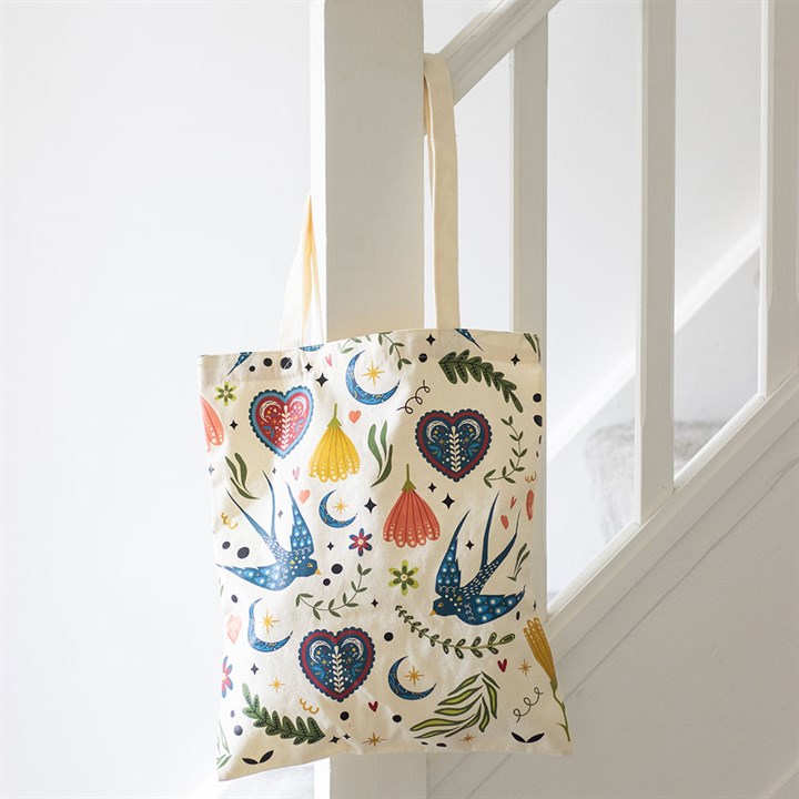 A cream polycotton tote bag adorned with colourful birds, flowers, hearts, leaves and crescent moons hangs from a white staircase bannister in a bright, minimalist space.
