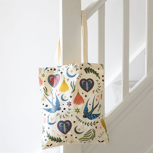 A cream polycotton tote bag adorned with colourful birds, flowers, hearts, leaves and crescent moons hangs from a white staircase bannister in a bright, minimalist space.