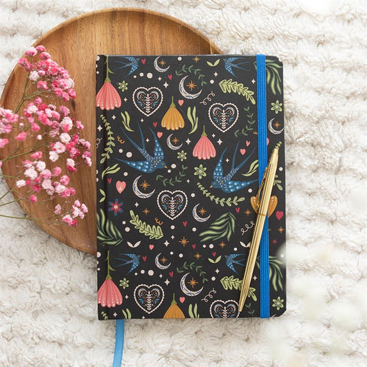A black A5 notebook with a colourful floral design and lined pages rests on a soft, white textured surface. A gold pen is placed on top, with pink flowers and a round wooden tray next to it.