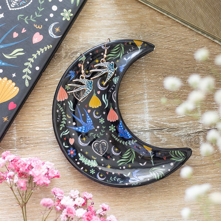 A crescent moon-shaped dish adorned with colourful floral and celestial patterns holds two bird-shaped earrings. Encircled by pink flowers and a sense of mystery, a floral notebook lies on a light wooden surface.