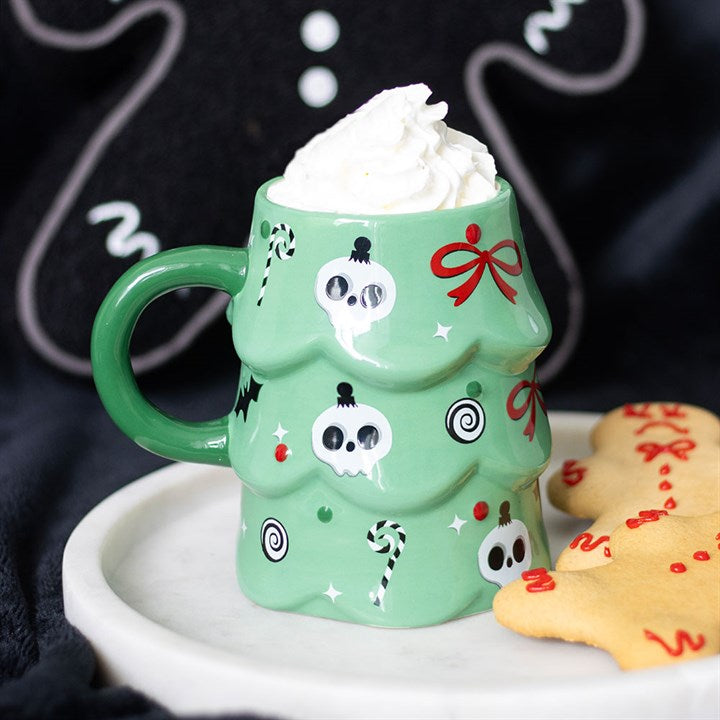 A green, spooky Christmas mug shaped like a tree, adorned with skull, candy cane and bow designs, overflows with whipped cream. It rests on a white plate next to decorated biscuits, set against a black gingerbread-figure background.