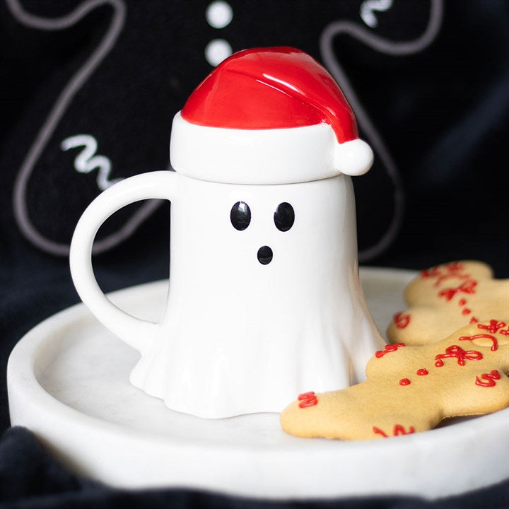 A ghost-shaped ceramic mug with black eyes and mouth, topped with a Father Christmas hat lid, sits on a white plate next to gingerbread biscuits. The black background features festive gingerbread man decorations.