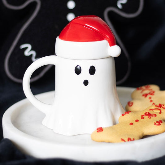 A ghost-shaped ceramic mug with black eyes and mouth, topped with a Father Christmas hat lid, sits on a white plate next to gingerbread biscuits. The black background features festive gingerbread man decorations.