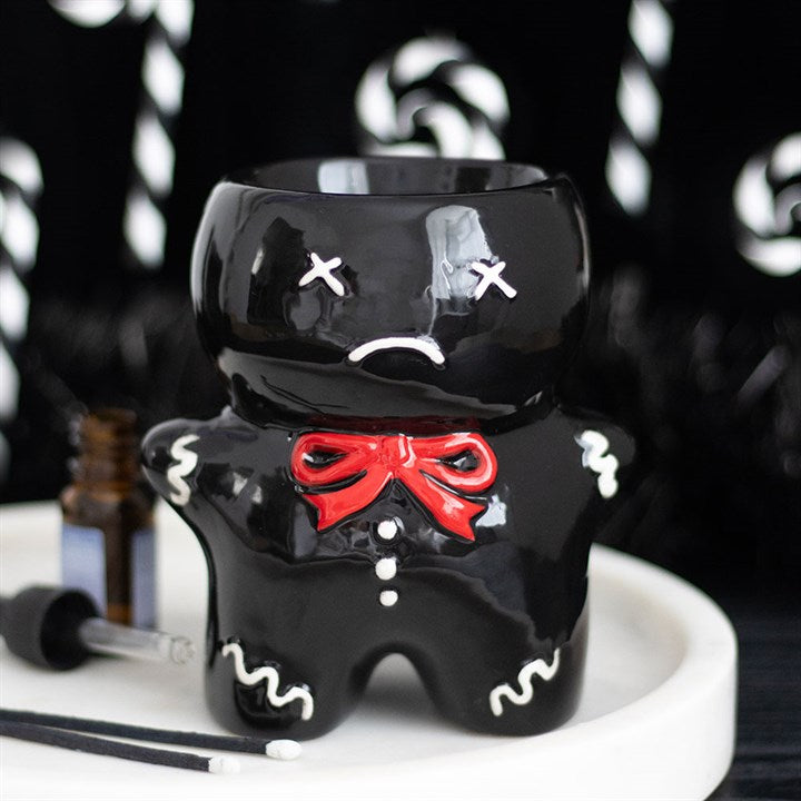 A glossy black mug shaped like a gingerdead man oil burner, featuring white ‘X’ eyes, a frown, red bow tie and icing details, stands on a plate—perfect as quirky festive décor. A dropper bottle and swirled background complete the scene.