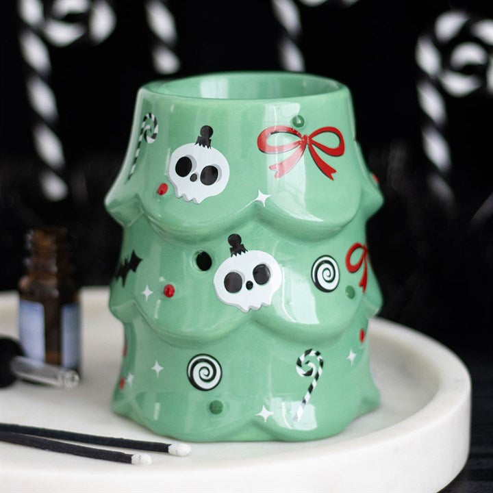 A green ceramic Christmas tree oil burner features white skulls, red bows, bats, candy canes, and swirl designs. This festive decoration sits on a white tray with an oil bottle and black-handled tools beside it.