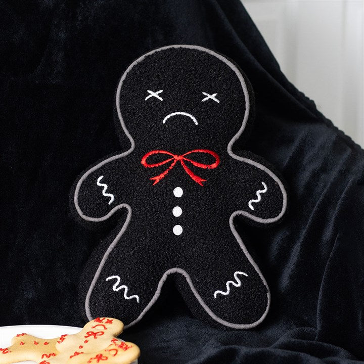 A quirky addition to your festive décor, this plush gingerdead man cushion features black fabric, white stitched detailing, a red bow, and a sad face. A biscuit-shaped gingerbread figure rests nearby on a black blanket.