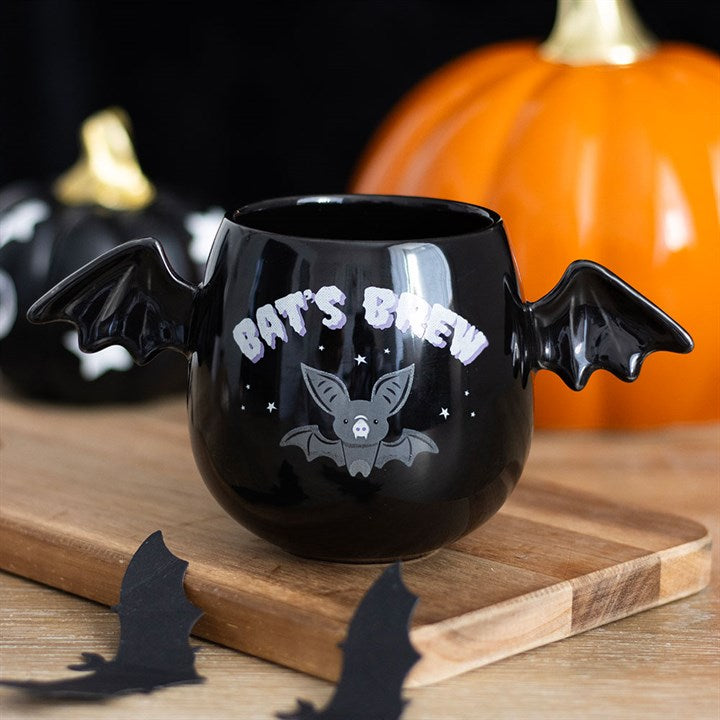 A ceramic mug shaped like a bat, complete with bat-wing handles, featuring the text Bats Brew and a cartoon bat. Orange and black pumpkins positioned behind it add a charming gothic touch to the Halloween theme.