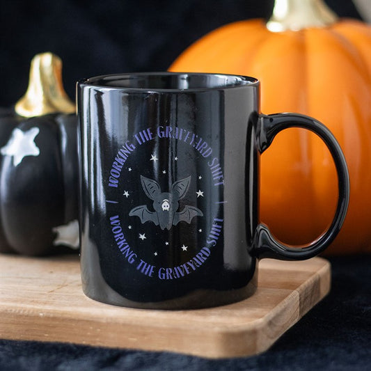 A ceramic mug featuring a bat and stars design sits on a wooden tray, ideal for night owls. “Working the Graveyard Shift” is printed in purple. Black and orange ceramic pumpkins complete the spooky style against a dark background.