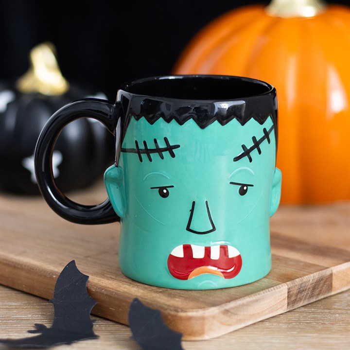 A grumpy monster mug shaped like Frankenstein’s creature, with green skin and black hair, sits on a wooden board surrounded by black and orange pumpkins and paper bats—a perfect quirky gift for Hallowe’en.
