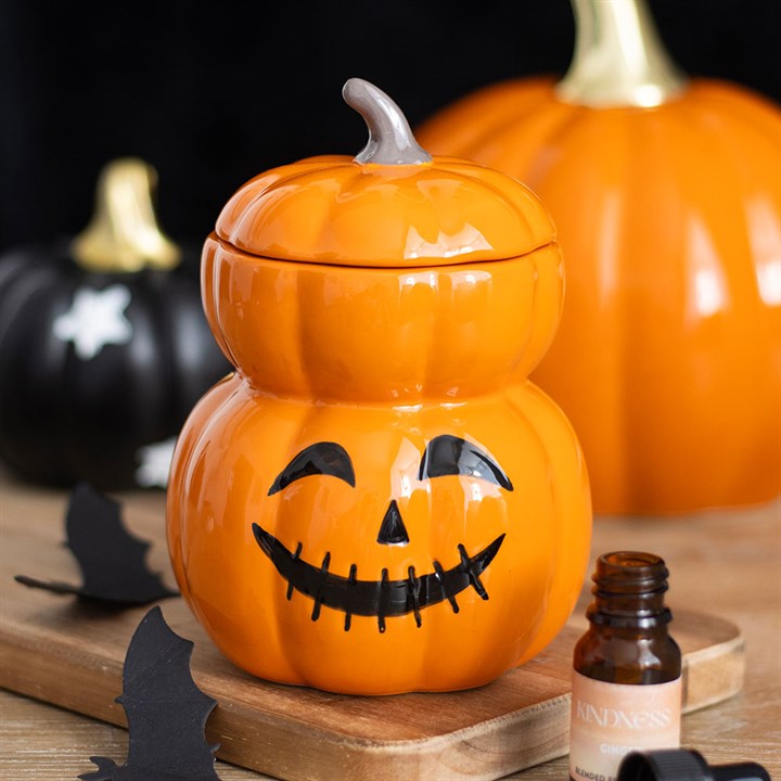 A ceramic pumpkin jar with a jack-o’-lantern face sits on a wooden tray, surrounded by black paper bats and small Halloween decorations. An amber glass bottle is in the foreground, with two pumpkins blurred in the background.