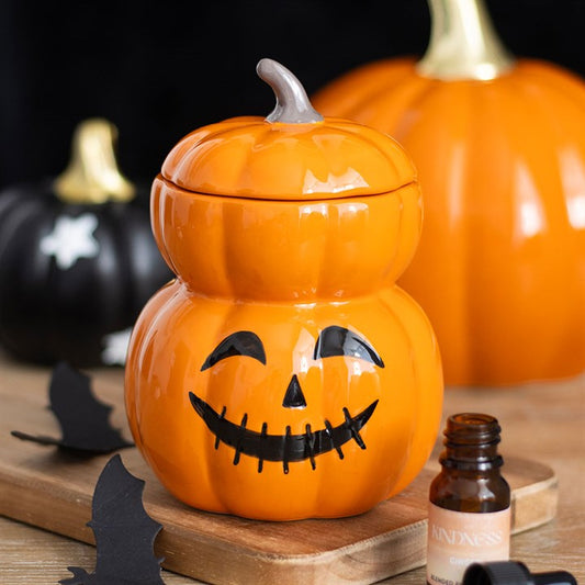 A ceramic pumpkin jar with a jack-o’-lantern face sits on a wooden tray, surrounded by black paper bats and small Halloween decorations. An amber glass bottle is in the foreground, with two pumpkins blurred in the background.