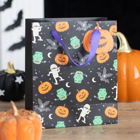 A Halloween-themed paper gift bag with purple handles features spooky icons such as jack-o’-lanterns, green monsters, bats, mummies, moons and stars. Real pumpkins and Halloween decorations sit on a wooden table nearby.