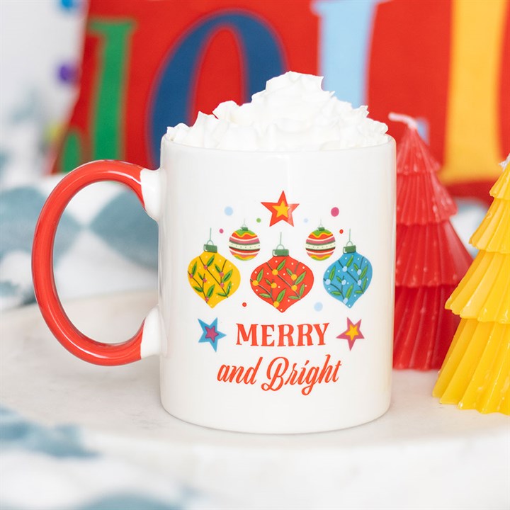 A festive Christmas mug with a red handle, decorated with colourful baubles and stars, bears the inscription Merry and Bright. The ceramic mug is topped with whipped cream and surrounded by red and yellow tree-shaped candles.