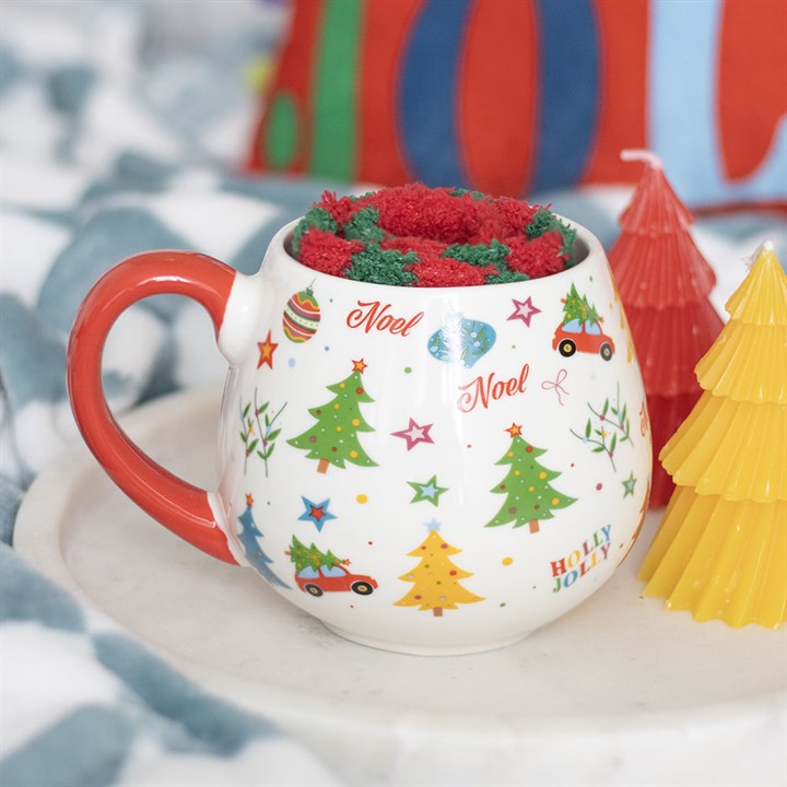 A cosy Christmas scene: a festive ceramic mug with a red handle, decorated with trees and holiday words, sits on a tray beside red and yellow Christmas tree-shaped candles—a perfect Christmas gift set.