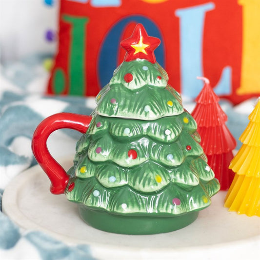 A ceramic Christmas tree mug adorned with colourful baubles, a red handle, and a red star on top sits on a white tray near cone-shaped candles, adding festive charm beside a cheerful holiday cushion.