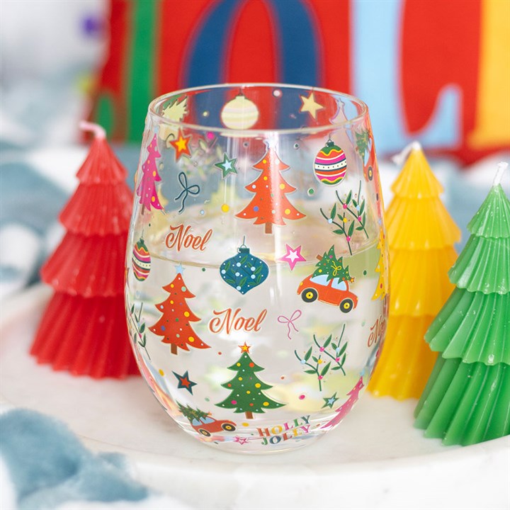 A festive stemless glass adorned with colourful Christmas designs—trees, baubles, stars, and the words “Noel” and “Holly Jolly.” It is surrounded by red, yellow and green tree-shaped candles on a white surface.