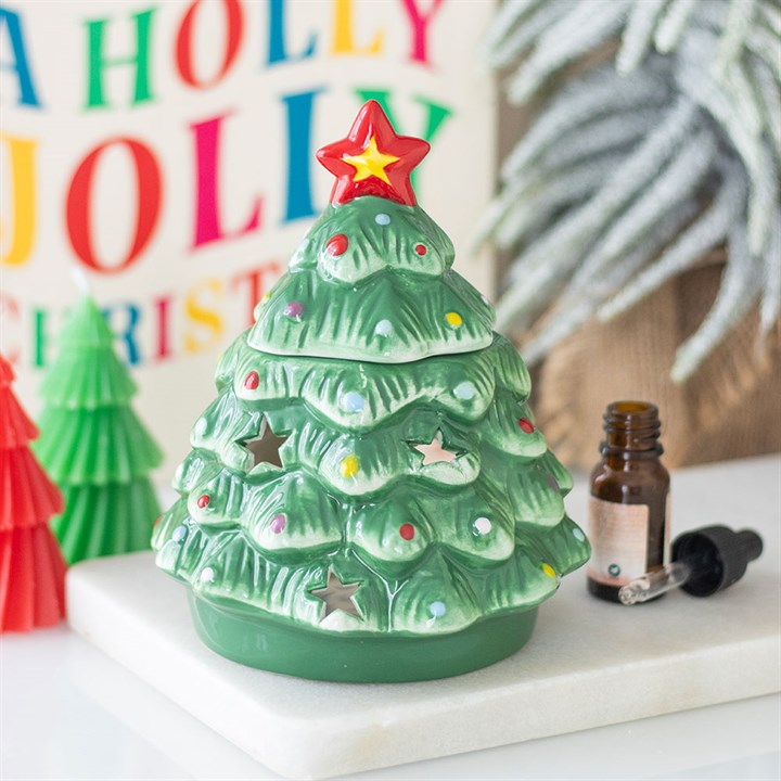 A ceramic oil burner shaped like a Christmas tree with star cut-outs and colourful dots sits on a marble tray beside an essential oil bottle, adding a festive touch alongside decorative trees and a holiday sign.