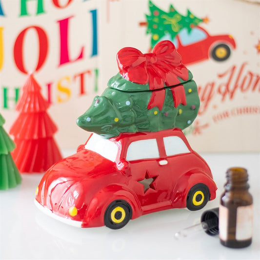 A ceramic red car with star-shaped cut-out windows and vintage charm carries a green Christmas tree with a red bow on its roof. Colourful festive decorations and a brown dropper bottle are in the blurred background.