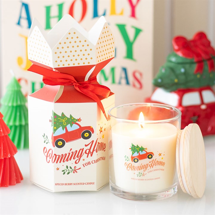 A spiced berries Christmas candle in a glass jar labelled ‘Coming Home for Christmas’, featuring a red car and tree design, sits beside a festive gift box amid cheerful holiday decorations and a colourful sign.