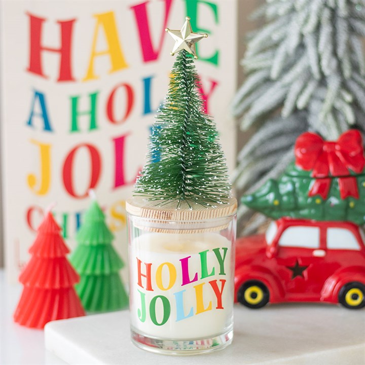 A festive candle with HOLLY JOLLY in colourful letters features a small bottle-brush Christmas tree and gold star. Behind it are a HAVE A HOLLY JOLLY CHRISTMAS sign, red toy car, and green and red tree decorations.