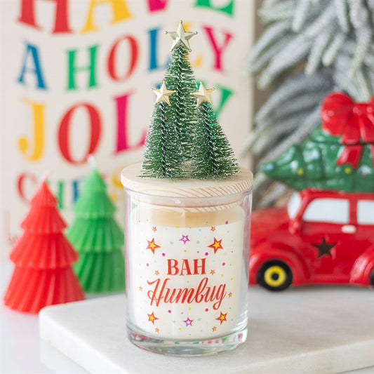 A Christmas candle labelled BAH Humbug with colourful stars sits on a table. Perfect as a stocking filler, it’s surrounded by red and green décor, a festive HAVE A HOLLY JOLLY sign, and cheerful Christmas trees on the lid.