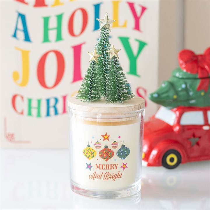 A candle in a glass jar featuring ‘Merry and Bright’ alongside Christmas ornament designs on the front. Its festive fragrance of cinnamon and spice is complemented perfectly by a lid adorned with three miniature trees topped with gold stars.