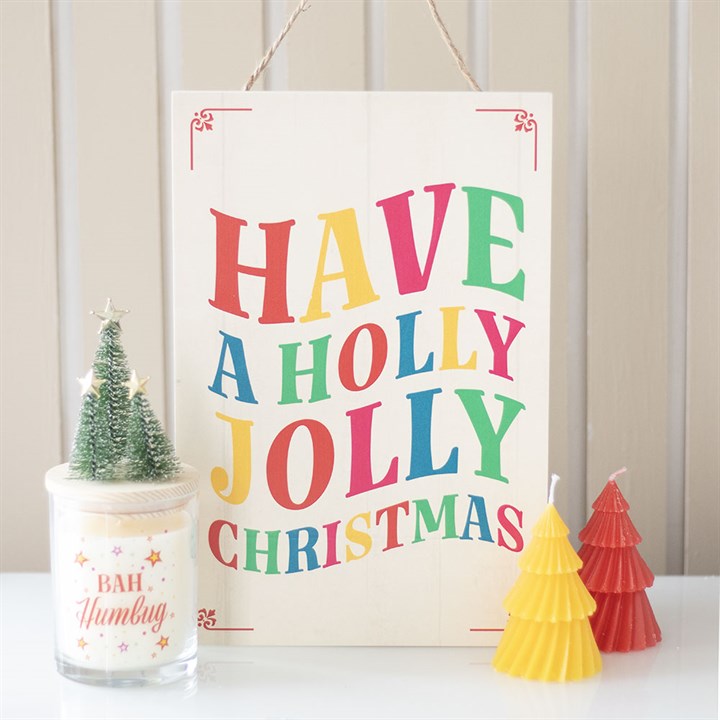 A festive hanging sign with colourful lettering reads Have a Holly Jolly Christmas, spreading Christmas cheer. In front are a small tree-shaped candle, a Bah Humbug candle, and two tree-shaped candles in yellow and red.