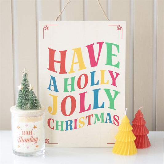 A festive hanging sign with colourful lettering reads Have a Holly Jolly Christmas, spreading Christmas cheer. In front are a small tree-shaped candle, a Bah Humbug candle, and two tree-shaped candles in yellow and red.