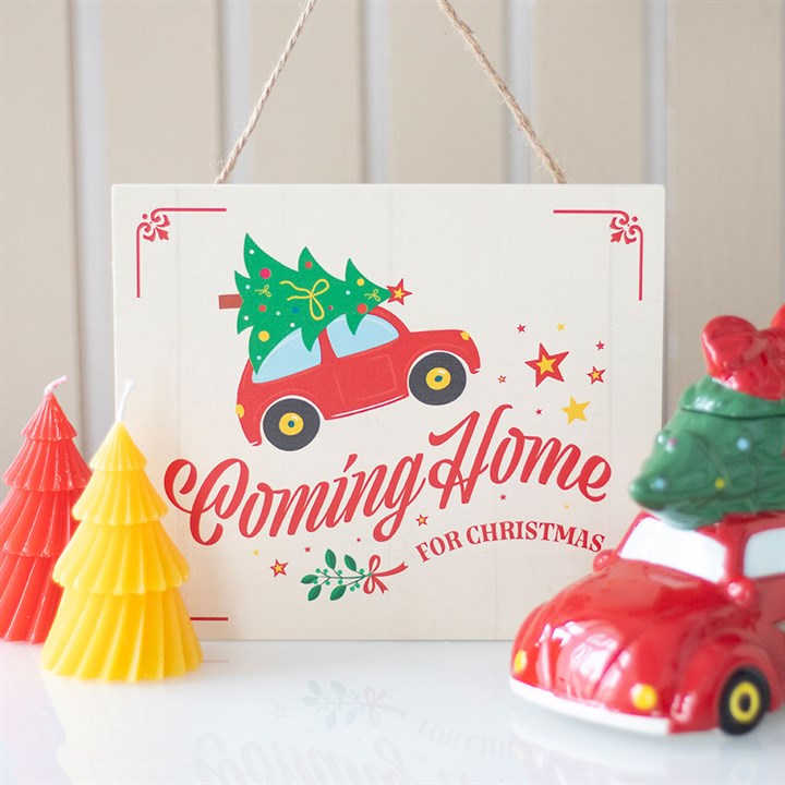 A festive Christmas sign reads “Coming Home for Christmas”, featuring a vintage red car carrying a Christmas tree. Red and yellow Christmas tree candles and a red car ornament are set against a light, striped background.