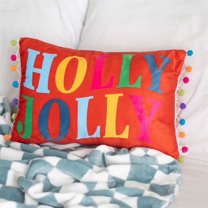 A festive cushion, this red rectangular pillow features colourful letters spelling HOLLY JOLLY and multicoloured pom-poms, adding cheerful seasonal décor to a white bed next to a blue and white geometric blanket.