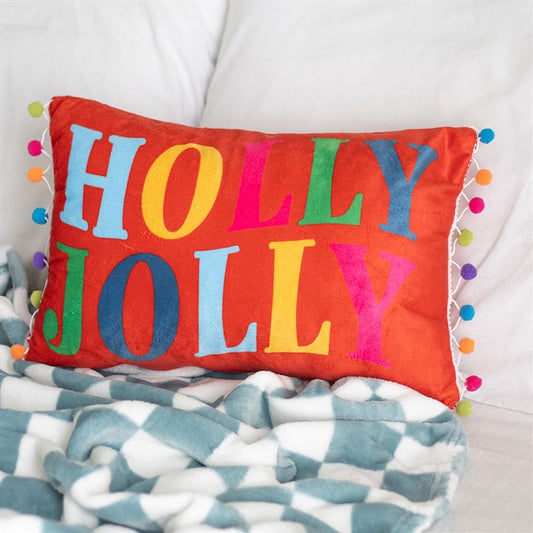 A festive cushion, this red rectangular pillow features colourful letters spelling HOLLY JOLLY and multicoloured pom-poms, adding cheerful seasonal décor to a white bed next to a blue and white geometric blanket.