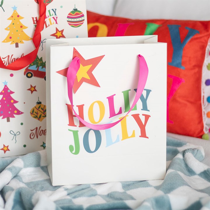 A white Holly Jolly gift bag with colourful letters and a pink ribbon handle sits on a blue and white blanket, surrounded by festive presents and a red Christmas cushion in the background.