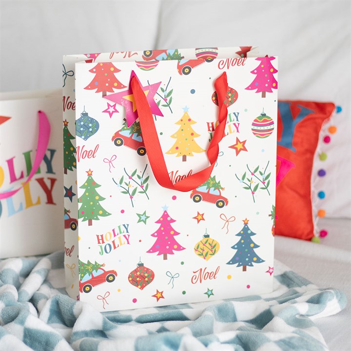 A Christmas gift bag with red handles features a festive design of colourful Christmas trees, baubles, stars, and nostalgic print reading Noel and Holly Jolly. It sits on a light blue and white patterned surface near some cushions.