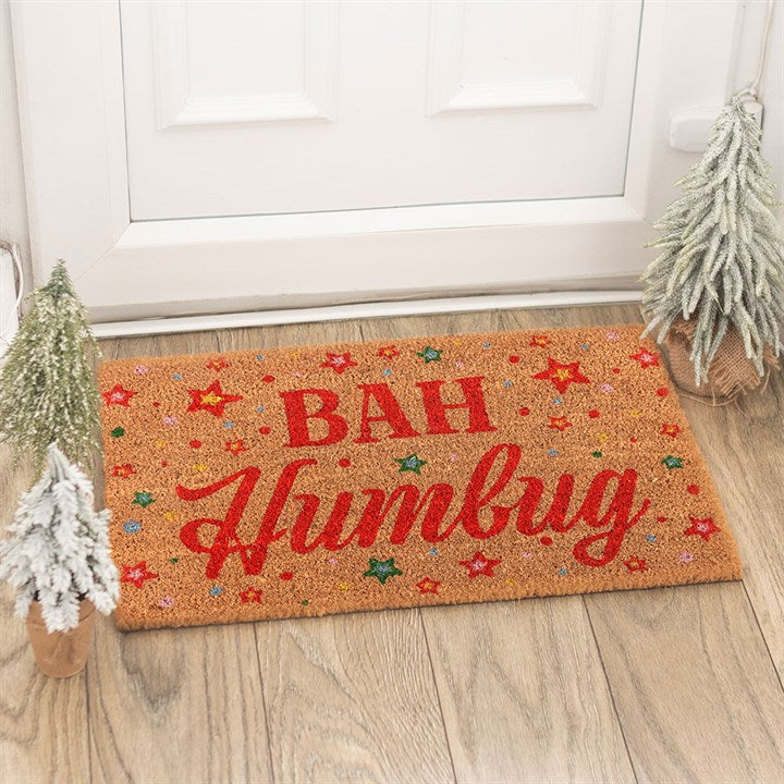 A ‘Bah Humbug’ doormat with red lettering and colourful stars sits in front of a white door, flanked by two small, snow-dusted artificial Christmas trees—a humorous doormat to spread festive cheer.