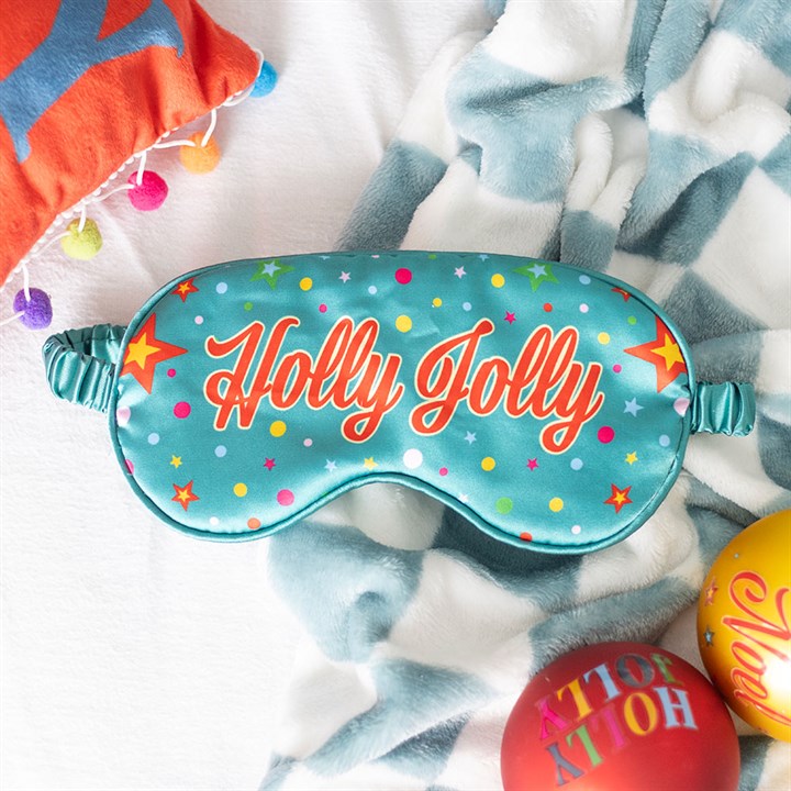A satin sleep mask with colourful stars and Holly Jolly in bold letters rests on a blue-and-white blanket, near decorations—a perfect Christmas stocking filler.