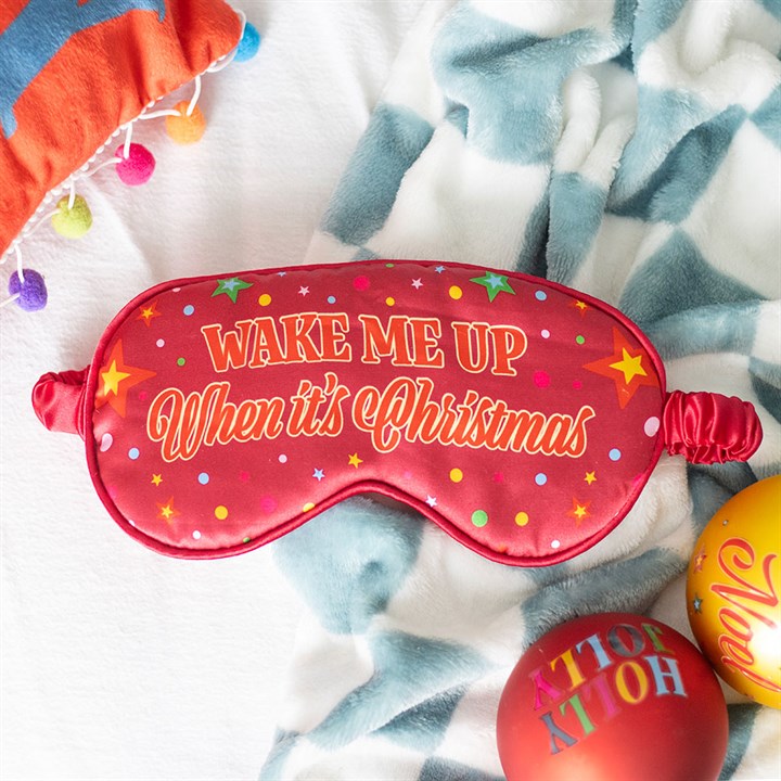 A red Christmas sleep mask with colourful stars and polka dots reads, “WAKE ME UP When it’s Christmas.” This festive gift rests on a soft white-and-blue blanket, with Christmas decorations nearby.