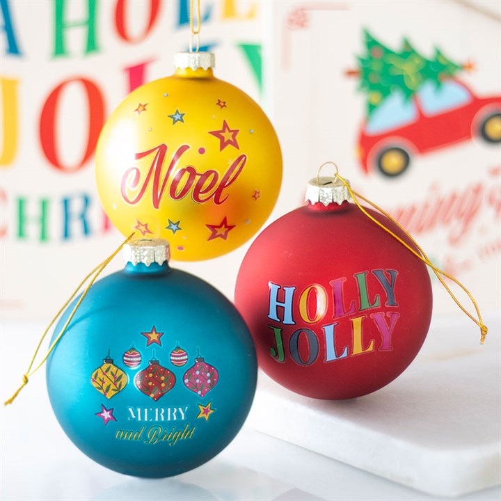 Three colourful glass Christmas baubles—yellow with Noel, red with Holly Jolly, and teal with Merry and Bright—bring festive cheer against a blurred holiday background featuring classic Christmas phrases.