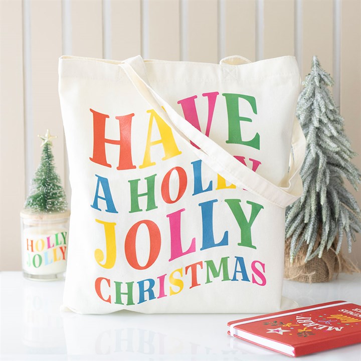A festive tote bag with rainbow-coloured text reading ‘Have a Holly Jolly Christmas’ sits on a white surface. Nearby are a Holly Jolly candle, a small frosted Christmas tree, and a red notebook adorned with gold stars.