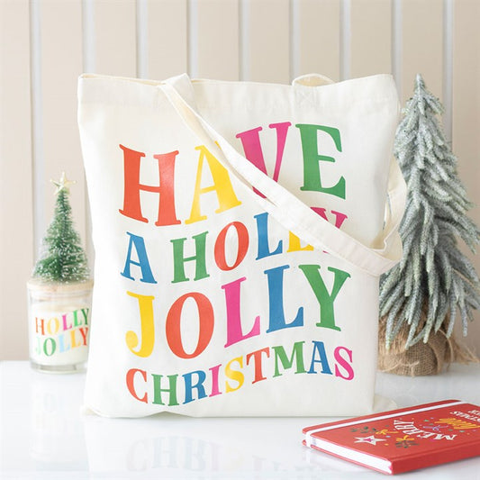 A festive tote bag with rainbow-coloured text reading ‘Have a Holly Jolly Christmas’ sits on a white surface. Nearby are a Holly Jolly candle, a small frosted Christmas tree, and a red notebook adorned with gold stars.