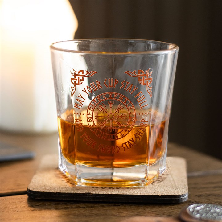 A glass of amber-coloured whisky rests on a Viking compass lowball glass atop a wooden table. The glass is adorned with Norse-inspired patterns and the phrase, May your cup stay full and your sword stay sharp.