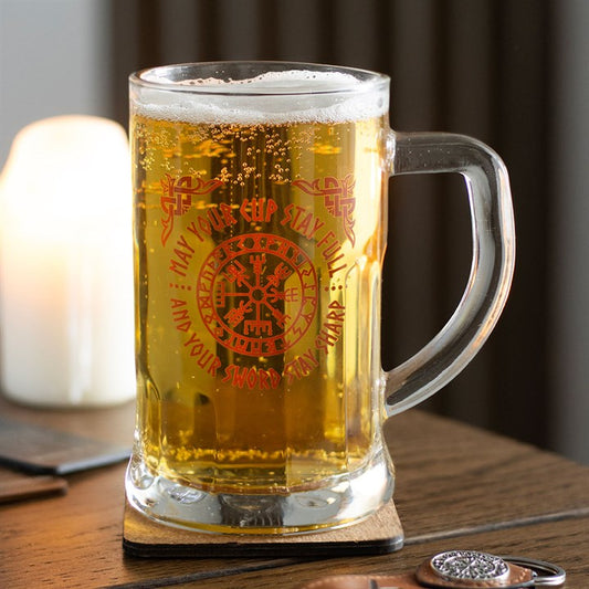 A clear glass Viking beer tankard brimming with golden ale rests on a wooden coaster, decorated with red Norse symbols. A lit candle and soft, blurred background evoke a cosy atmosphere—an ideal Norse mythology gift.