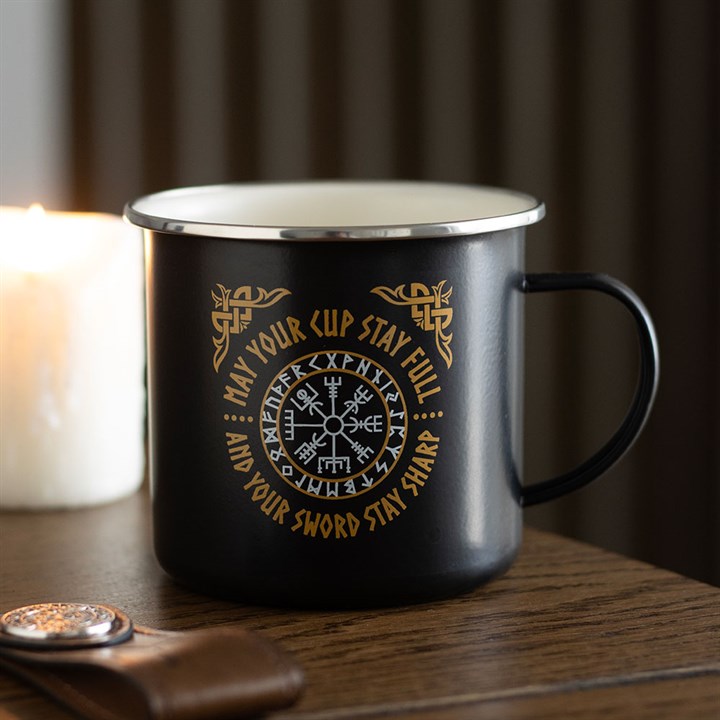 A black enamel-style Viking mug with gold Vegvisir compass designs rests on a wooden table. The mug reads, “May your cup stay full and your sword stay sharp,” encircling a Norse symbol. A candle and leather keyring are nearby.
