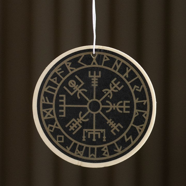 A round, black Viking Compass air freshener adorned with tan Norse runes and a Vegvisir symbol dangles from a white string, emitting an earthy scent against a blurred, dark brown backdrop.