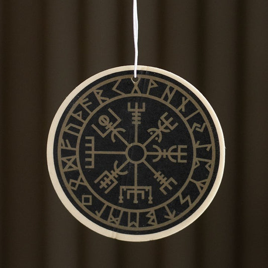 A round, black Viking Compass air freshener adorned with tan Norse runes and a Vegvisir symbol dangles from a white string, emitting an earthy scent against a blurred, dark brown backdrop.