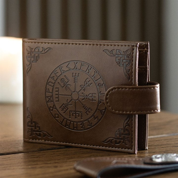 A brown leather Viking Compass wallet with a snap fastening, embossed with Norse symbols and a circular Vegvisir motif, rests on a wooden surface with a blurred candle in the background.